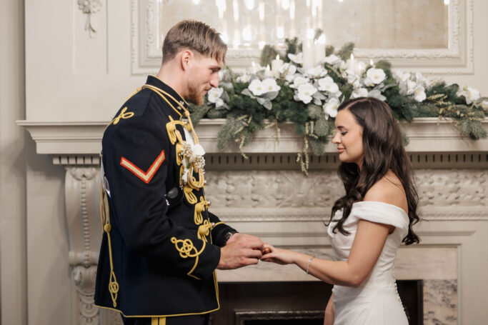 A man in a formal military uniform places a ring on the finger of a woman in an off-the-shoulder white wedding dress, standing together in front of a decorated fireplace with white flowers and greenery.