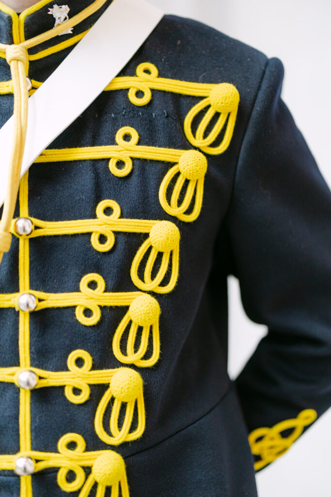 A close-up of a dark navy or black military-style uniform with ornate yellow braided trim and decorative buttons, showing detailed cords and knots on the chest and sleeve.