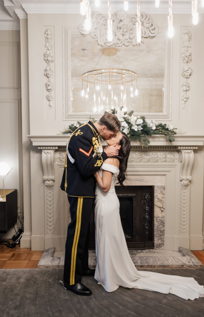 A couple in formal attire, the man in a military uniform and the woman in a white dress, share a kiss in front of an ornate fireplace decorated with greenery and flowers.