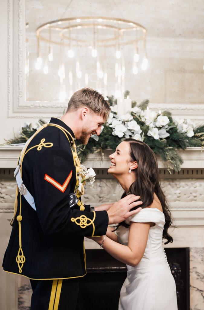 A smiling groom in a formal military uniform and a bride in an off-the-shoulder white dress laugh together in front of a decorated fireplace with white flowers and greenery. A chandelier hangs in the background.