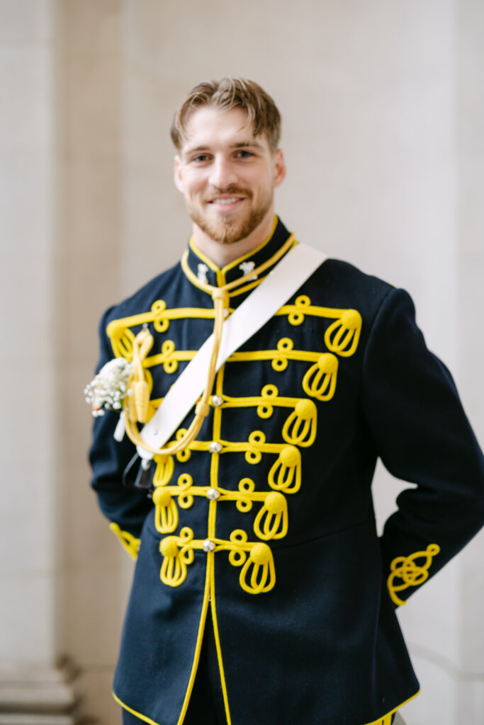 A man wearing a formal black uniform with bright yellow trim and decorative buttons stands smiling, with a small white boutonniere on his chest and his hands behind his back, in front of a light-colored stone wall.