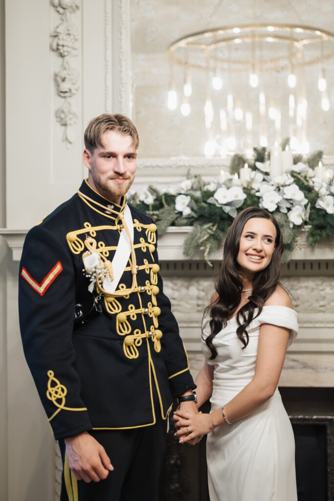A man in a formal military uniform and a woman in an off-the-shoulder white gown stand holding hands and smiling, in front of a fireplace decorated with white flowers and greenery. A chandelier hangs overhead.