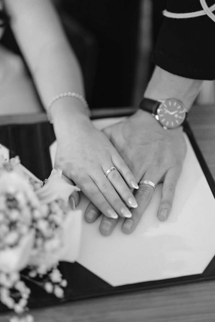 A black and white photo of two hands with wedding rings resting on a surface, one hand gently placed over the other. A bouquet of flowers is visible in the foreground.