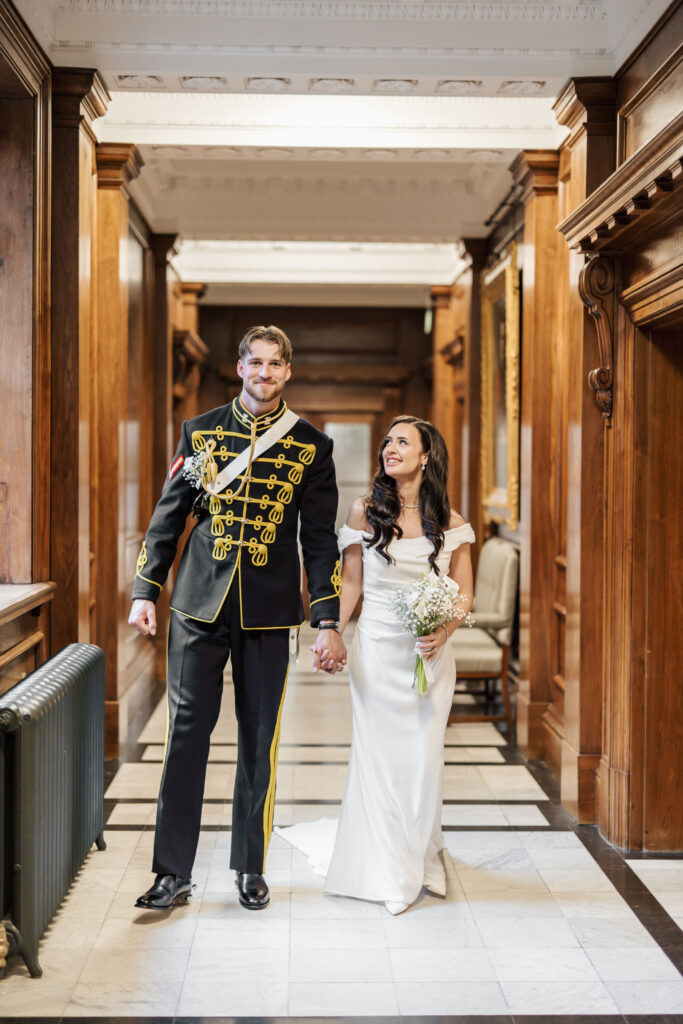 A smiling couple walks hand in hand in a grand hallway; the man wears a formal military uniform, and the woman wears a white wedding dress, holding a bouquet of flowers.