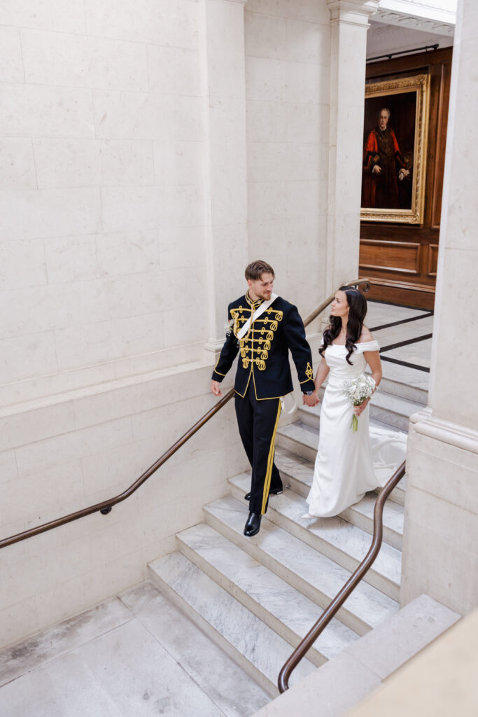 A bride in a white dress and a groom in a black and gold military-style uniform walk up marble stairs, holding hands, in a grand building with white walls and a portrait hanging on the wall.