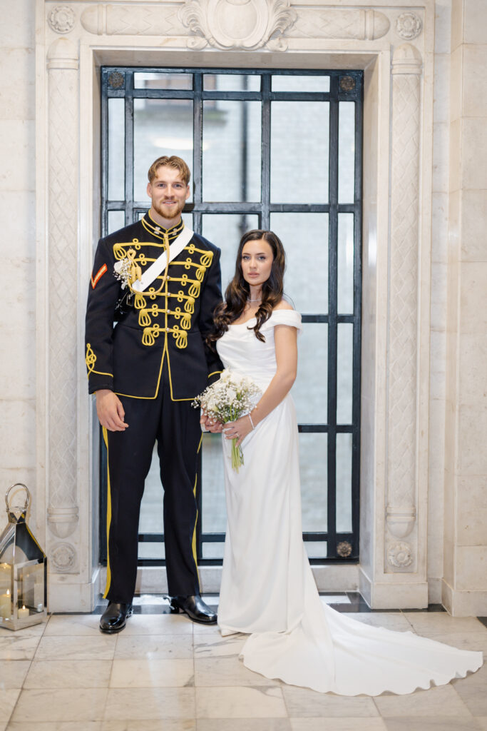 A couple stands side by side indoors; the man wears a formal military uniform with gold details, and the woman wears an off-shoulder white wedding gown, holding a bouquet of flowers. They pose in front of a decorative gate.