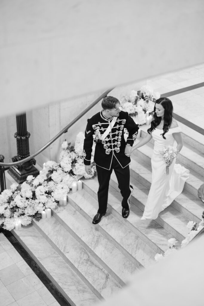 A bride in a white gown and a groom in a formal military uniform walk down marble stairs decorated with flowers and candles, smiling at each other. The scene is elegant and romantic in black and white.
