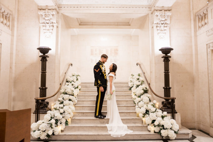 A bride in a white dress and a groom in a military uniform stand on elegant marble stairs decorated with white flowers and candles, gazing at each other in a grand, ornate hall.