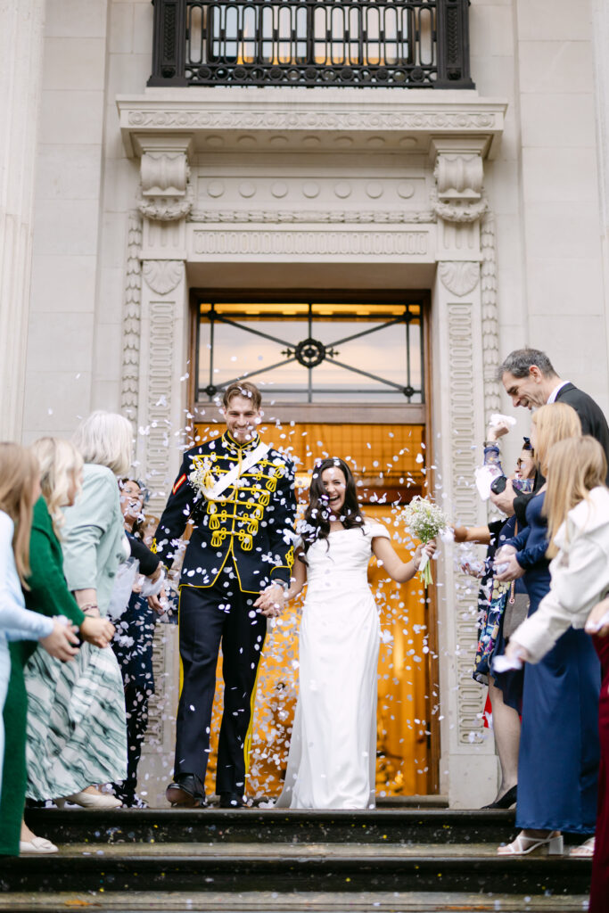 A newlywed couple, the groom in a decorated military uniform and the bride in a white dress, walk down steps as guests throw confetti and celebrate outside an ornate building.