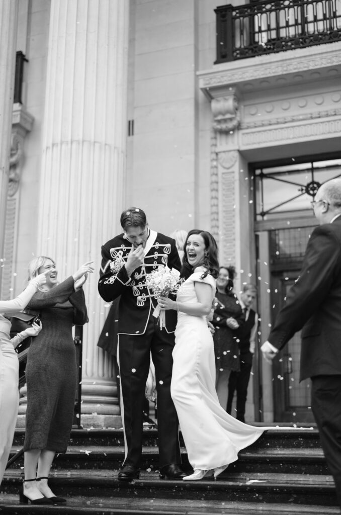A bride and groom, smiling and laughing, walk down the steps of a grand building as guests throw confetti. The groom is in a decorated uniform, and the bride wears a white dress. Others celebrate around them.