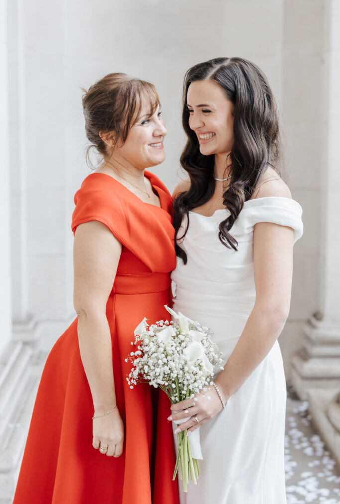 Two women stand close together, smiling warmly at each other. One wears a white dress and holds a bouquet of white flowers, while the other wears a bright red dress. They appear happy and are in an elegant, light-colored setting.