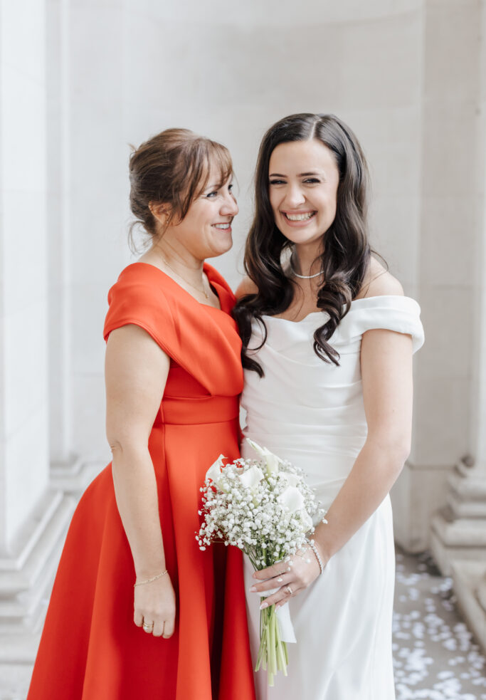Two women pose happily together in elegant dresses; one wears white and holds a bouquet, while the other wears a bright red dress. They stand close, smiling in a light-filled, columned setting.