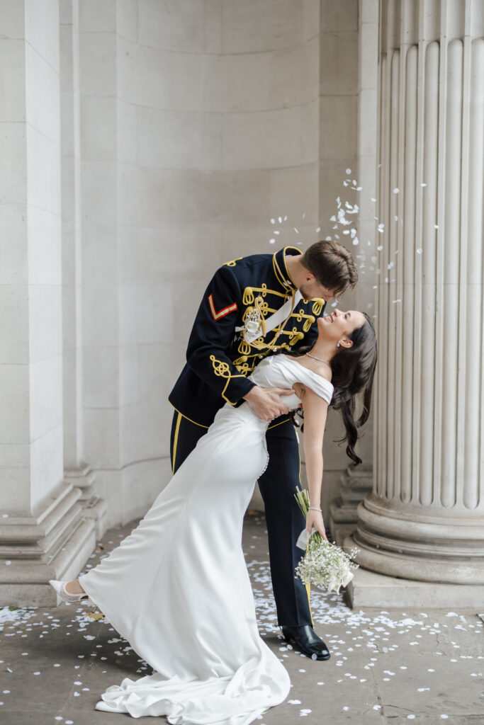 A groom in a military uniform dips and kisses his bride, who wears a white dress and holds a bouquet, as white confetti falls around them in front of tall stone columns.