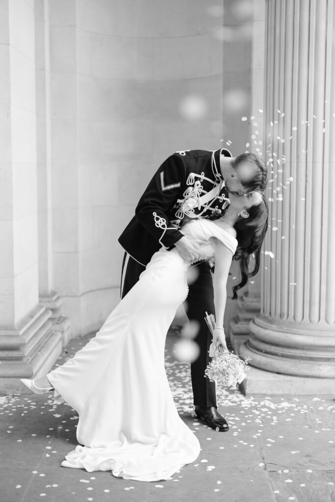 A groom in a formal military uniform dips and kisses his bride, who wears a long white dress and holds a bouquet, as flower petals fall around them in front of stone columns.