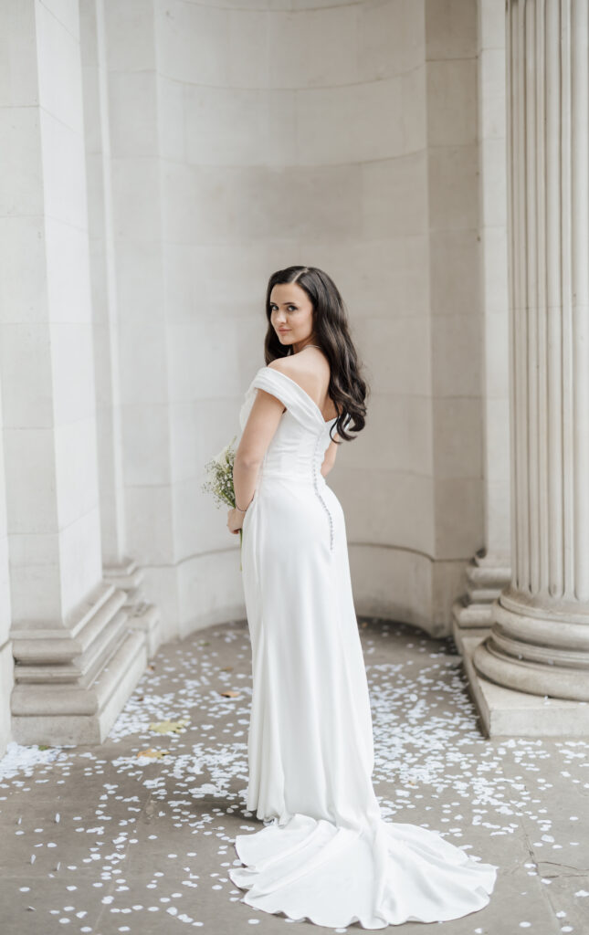 A bride with long dark hair in an off-the-shoulder white gown stands between stone columns, holding a bouquet and looking over her shoulder. White flower petals are scattered on the ground.