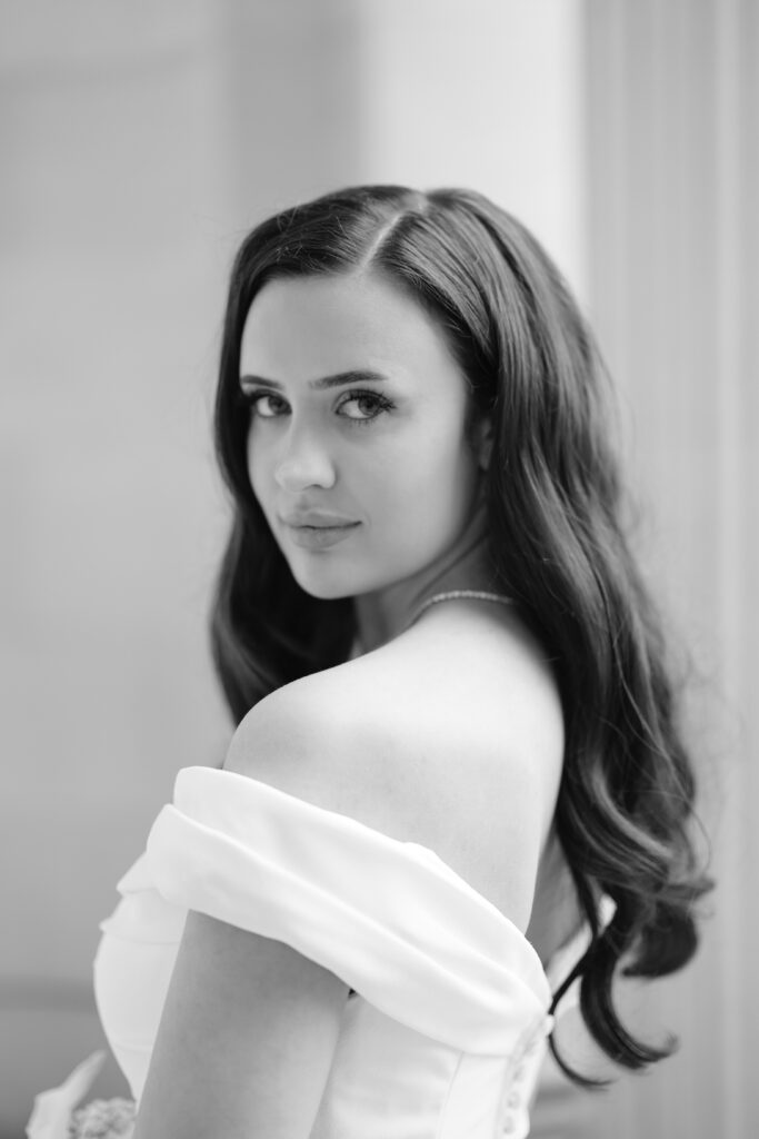 Black and white portrait of a woman with long, wavy hair wearing an off-the-shoulder dress, looking over her shoulder toward the camera with a soft, neutral expression.