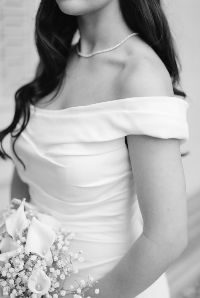A black and white photo of a woman in an off-the-shoulder wedding dress holding a bouquet of calla lilies and babys breath, with long wavy hair and a simple necklace.