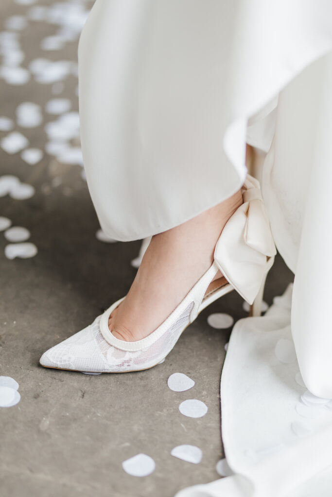 A close-up of a person wearing a white lace high heel shoe with a large bow, standing on a floor scattered with white confetti. The person is dressed in a flowing white garment, possibly a wedding dress.