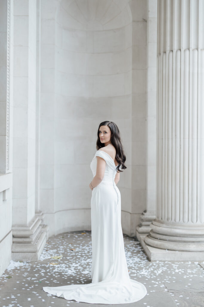 A woman in a white, off-the-shoulder wedding dress stands among tall stone columns, looking over her shoulder. White petals are scattered on the ground around her. The setting is bright and elegant.
