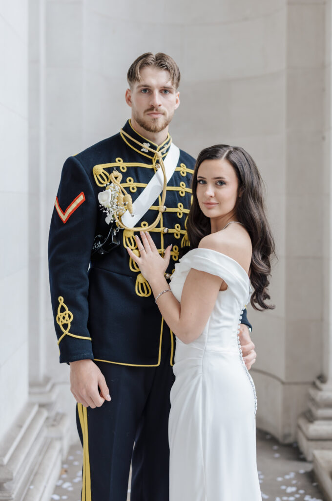 A man in a formal military dress uniform stands beside a woman in an off-shoulder white gown. They are posing together in an elegant, columned setting, both looking toward the camera.