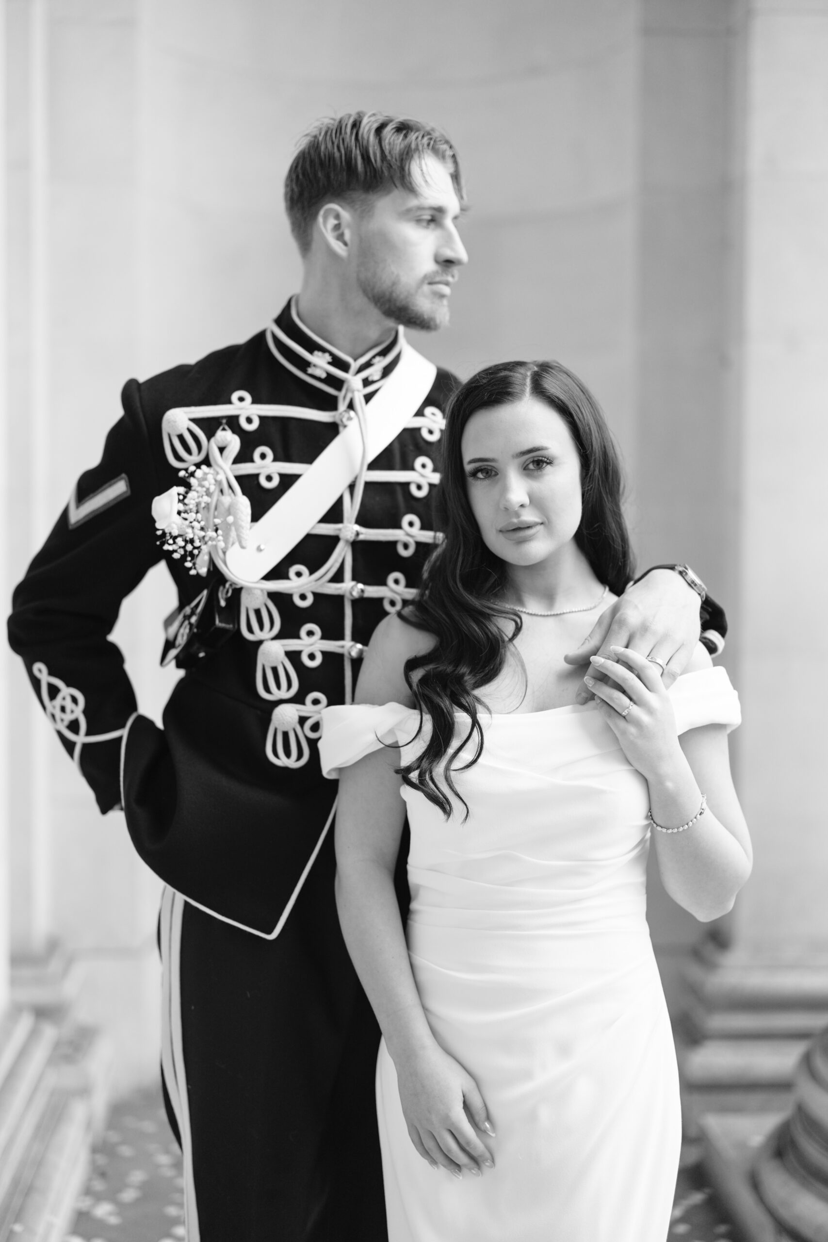 A man in a formal military uniform stands beside a woman in an off-the-shoulder white dress. The man rests his hand on her shoulder while she holds his hand, both looking in different directions, in a softly lit setting.