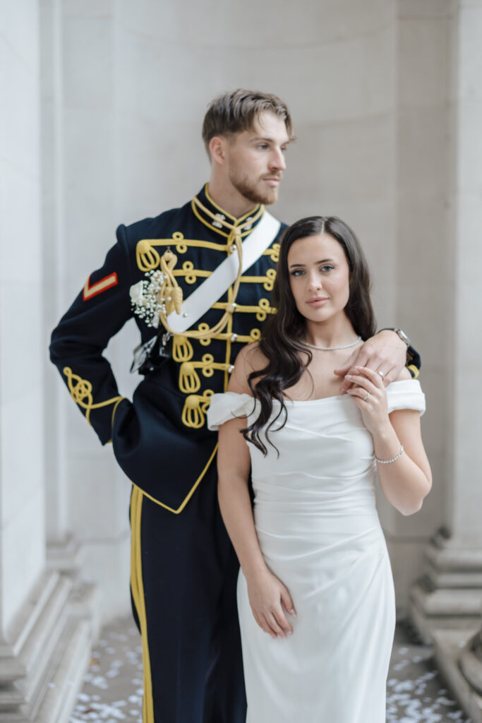 A groom in a military dress uniform stands with his arm around a bride in an off-the-shoulder white gown, both posing for a portrait in an elegant, marble-columned hallway.