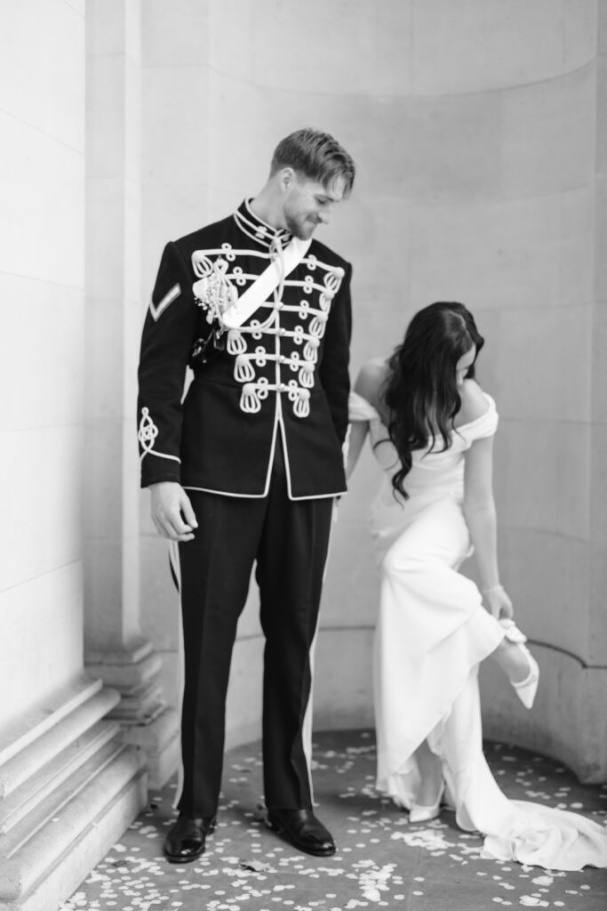 A man in a formal military uniform stands beside a woman in a white wedding dress who is adjusting her shoe. They are standing against a stone wall with flower petals scattered on the ground.