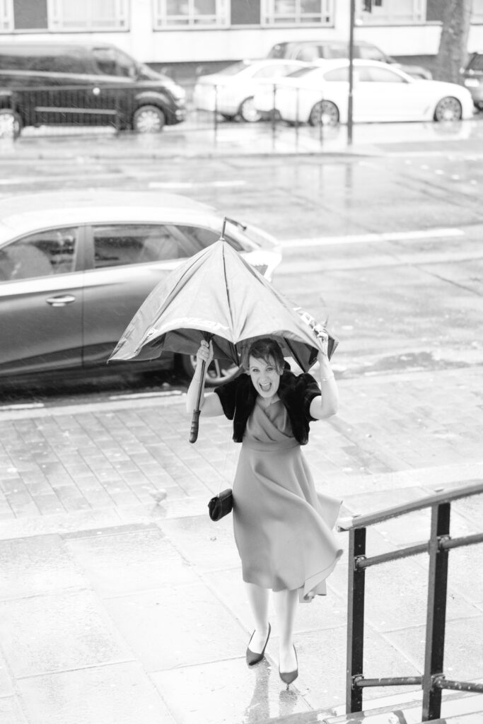 A woman in a dress smiles as she runs up stairs in the rain, holding a partially collapsed umbrella. Wet pavement and parked cars are visible in the background. The image is in black and white.