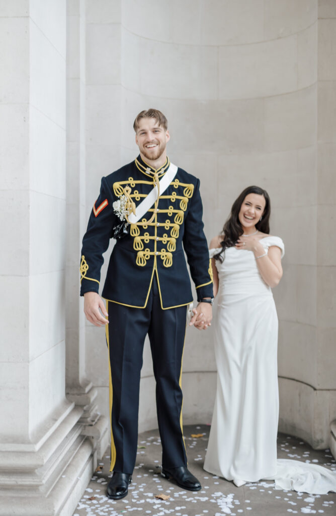 A smiling groom in a formal military uniform stands holding hands with a bride in a white gown. They are surrounded by white stone walls and scattered flower petals on the ground.