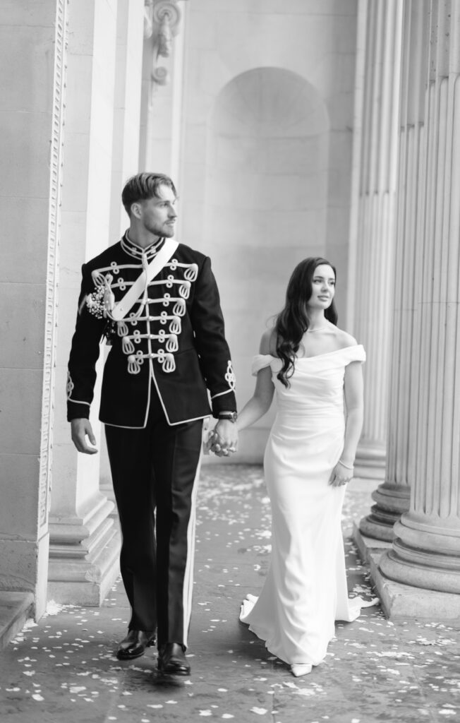 A man in a formal military-style uniform and a woman in an off-shoulder white gown walk hand in hand between tall stone columns, with flower petals scattered on the ground.