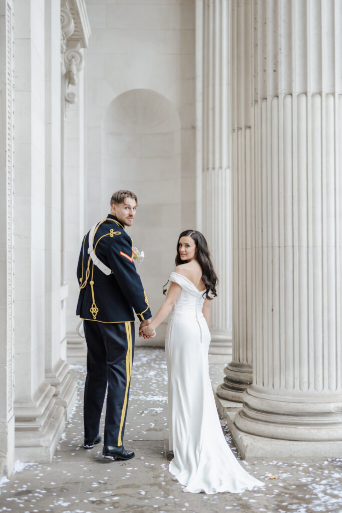 A bride in a white gown and a groom in a formal military uniform hold hands and look back while standing between tall white columns, with white flower petals scattered on the ground.