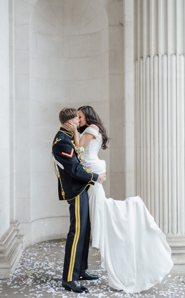 A groom in a formal military uniform lifts and kisses a bride in a white gown, surrounded by scattered white confetti in an elegant, columned setting.