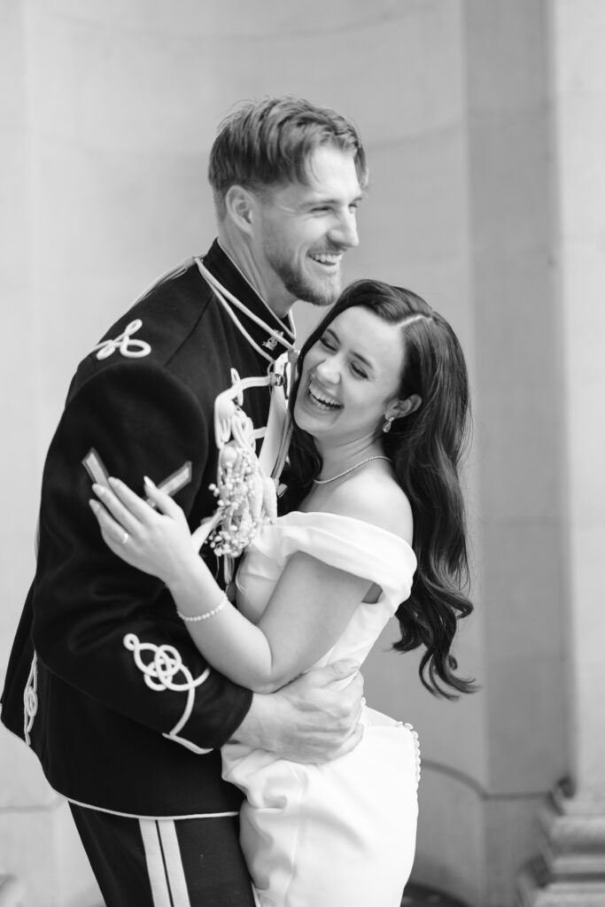 A man in a formal military uniform embraces a woman in an off-the-shoulder white dress. Both are laughing joyfully, sharing a happy moment in front of a light stone wall. The photo is in black and white.