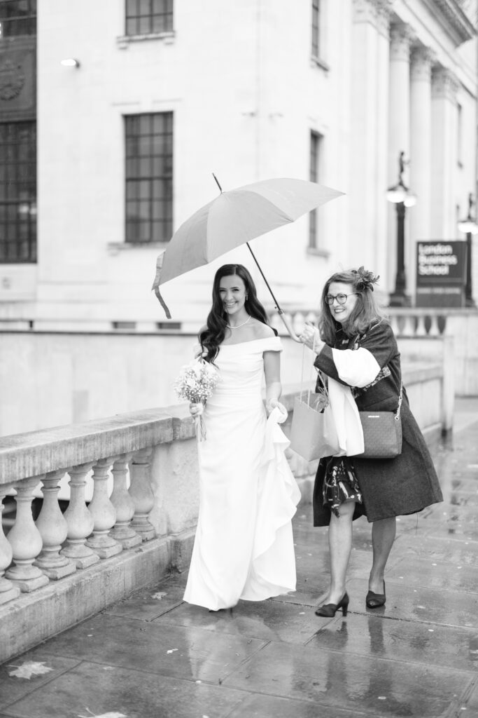 A bride in a white gown walks outside on a rainy day, holding her dress and bouquet, while another woman beside her holds a large umbrella over them. Both are smiling. The scene is in front of grand stone buildings.