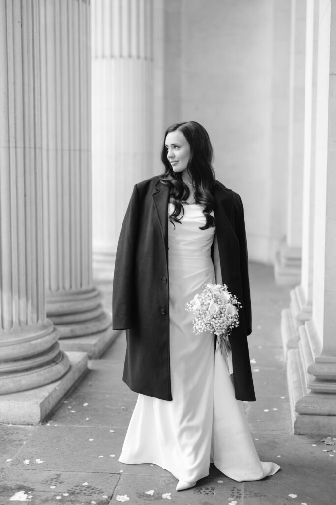A bride in a white dress and dark coat holds a bouquet, standing between tall columns. She looks to the side, smiling softly. The image is in black and white.