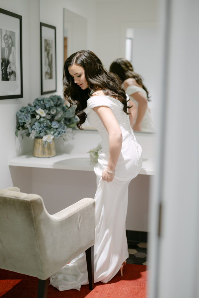 A bride in a white dress stands by a vanity, adjusting her gown. She has long, dark hair and is looking down. The room is softly lit and decorated with flowers and framed photos on the wall.