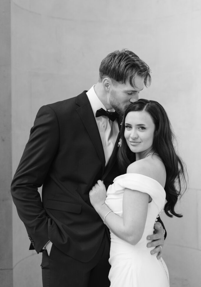 A man in a suit gently kisses the forehead of a woman in an off-the-shoulder wedding dress. They are embracing, and the woman smiles softly at the camera. The photo is in black and white.