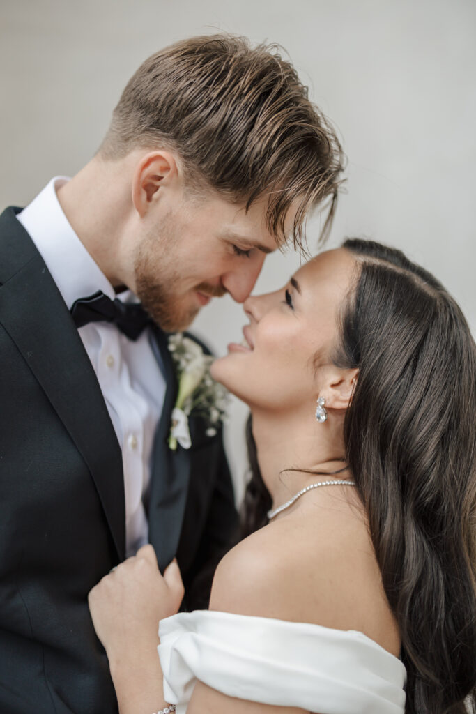 A bride and groom in formal wedding attire lovingly embrace and smile at each other, their foreheads touching. The groom wears a black tuxedo and the bride wears an off-the-shoulder white dress.