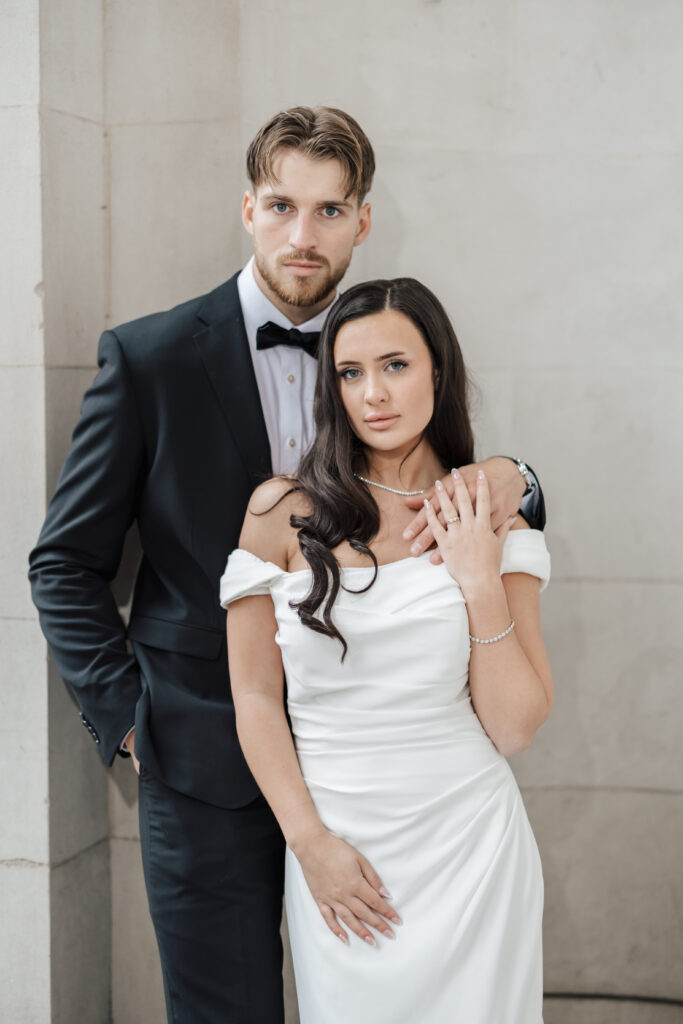 A man in a black tuxedo stands beside a woman in an off-the-shoulder white dress. The man rests his hand on her shoulder as they both look at the camera against a light stone wall background.