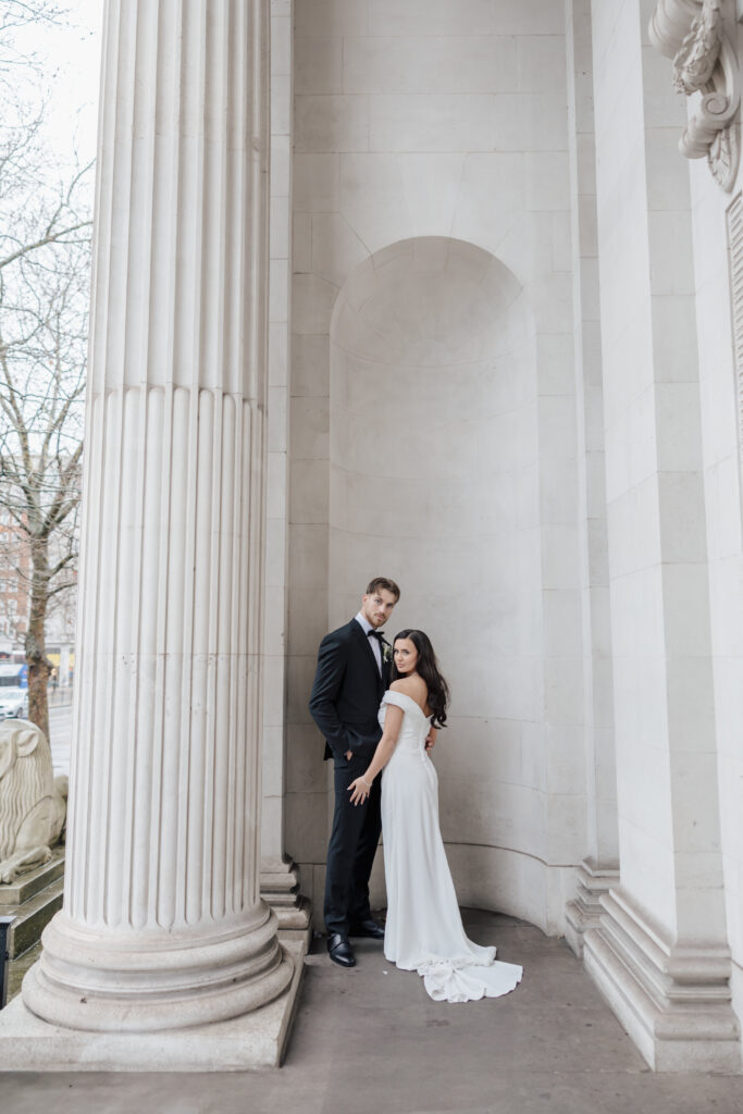 A bride in a white dress and a groom in a dark suit stand close together between tall marble columns and an arched alcove, posing for a wedding photo outside a grand building.