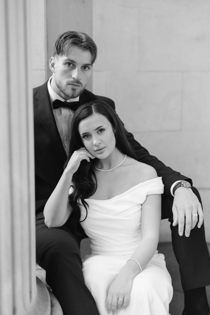 A man in a black tuxedo and a woman in an off-shoulder white dress sit closely together, posing for a formal portrait. The woman is seated and leaning against the man, who has his arm around her. They both have calm, serious expressions.