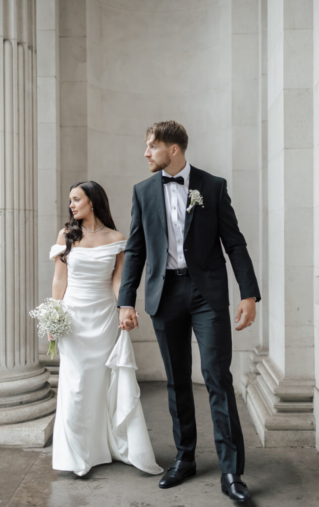 A bride in an off-the-shoulder white gown holds a bouquet and a groom in a black tuxedo walk hand-in-hand beside tall stone columns, both looking away from the camera.