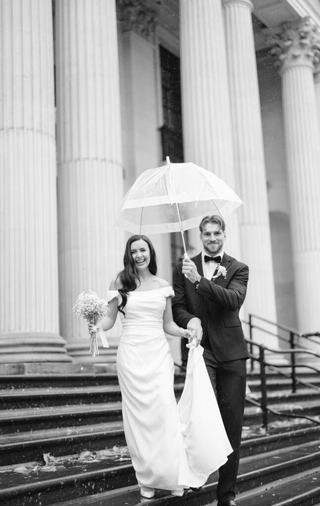 A bride and groom walk down stone steps in front of a columned building. The groom holds a clear umbrella over them as it rains. The bride smiles, holding a bouquet and lifting her dress. The photo is in black and white.