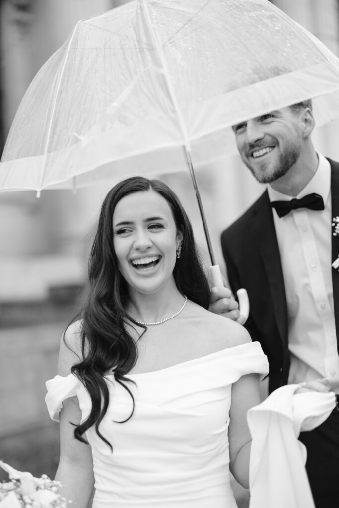 A bride in an off-shoulder white dress smiles while a groom in a tuxedo holds a clear umbrella above them. Both appear happy and are outdoors. The image is black and white.