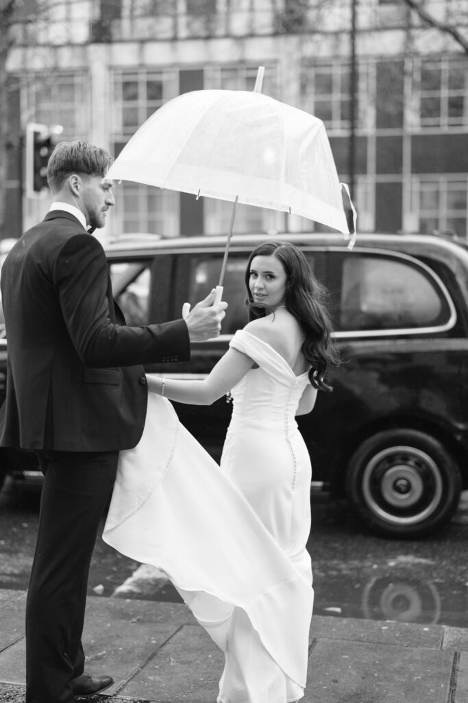 A man in a suit holds an umbrella over a woman in a white wedding dress as they stand on a wet city street near a black taxi. The woman looks back over her shoulder, holding her dress.