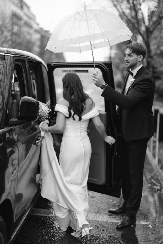 A groom holds an umbrella for a bride as she steps into a car on a rainy day. The bride wears a fitted, off-the-shoulder gown and holds up her dress, while the groom is dressed in a suit. The scene is in black and white.