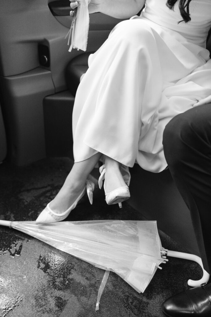 A bride in a white dress and heels sits with her legs crossed inside a vehicle. A clear umbrella lies on the wet floor, and part of another person in dark clothing is beside her.