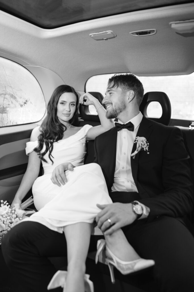 A newlywed couple dressed in a wedding gown and suit sit relaxed in the backseat of a car, smiling at each other. The bride leans against the seat with her legs across the groom’s lap. Black and white photo.