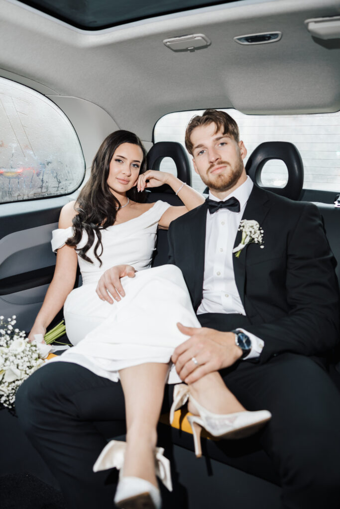 A bride in an off-shoulder white dress and a groom in a black tuxedo sit together in the back seat of a car, both looking confidently at the camera. The bride holds a bouquet of white flowers.