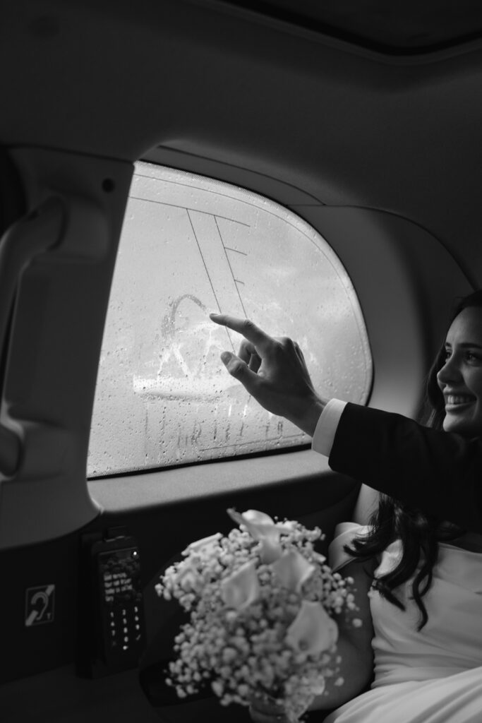 A bride, smiling in a car, draws a heart on a foggy window with her finger while holding a bouquet of flowers in her lap.